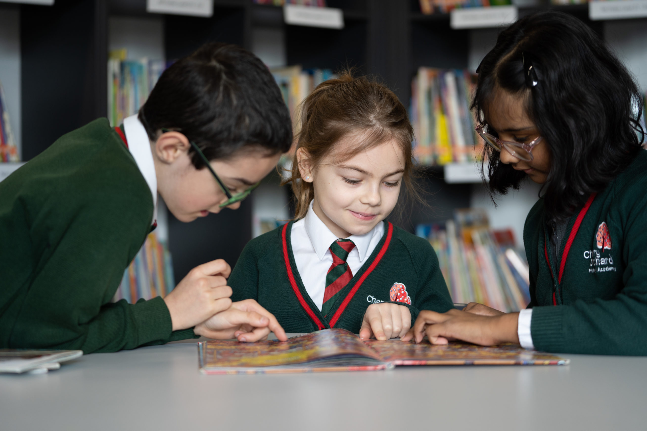 Three pupils, one boy and two girls, are pictured sat together at a desk reading a storybook and smiling.